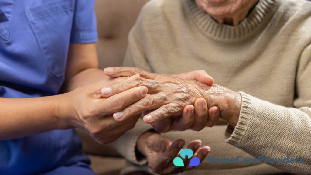 a tender moment where a caregiver in a blue uniform is gently holding the hands of an elderly person wearing a beige sweater (1)